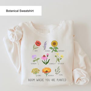 May include: Cream-colored botanical sweatshirt featuring colorful illustrations of various flowers, including poppy, lavender, and sunflower. The text "BLOOM WHERE YOU ARE PLANTED" is printed at the bottom, with the words "Botanical Sweatshirt" at the top.