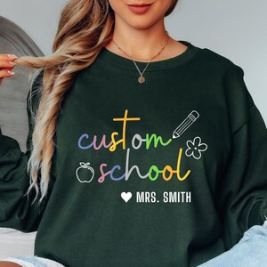 Dark green custom teacher sweatshirt featuring rainbow text “custom school” with apple, pencil, and flower icons, personalized with teacher’s name. Cozy unisex crewneck perfect for teacher appreciation gifts, back-to-school outfits, or school spirit day apparel