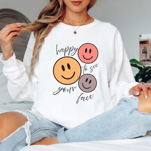 May include: White sweatshirt with a graphic of three smiley faces in orange, pink, and gray, with the words "happy to see your face" in black script. The sweatshirt is a casual, comfortable style.