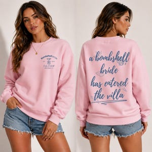 May include: A light pink long-sleeve sweatshirt featuring a small blue graphic on the front with the text Bombshell Bride Est 2023 Emily's Bachelorette Miami Beach, FL. The back displays the blue text a bombshell bride has entered the villa.
