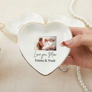 May include: A heart-shaped, white ceramic trinket dish with a gold rim. The dish features a printed photograph of a mother and child, with the text "Love you, Mom Emma & Noah". A pearl necklace and other jewelry are visible.