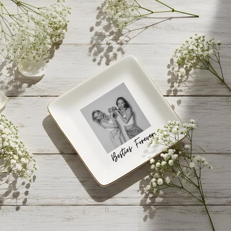 May include: Square white ceramic trinket dish with a gold rim, featuring a black and white photo of two people and the text "Besties Forever". The dish is surrounded by white flowers on a light wood surface.