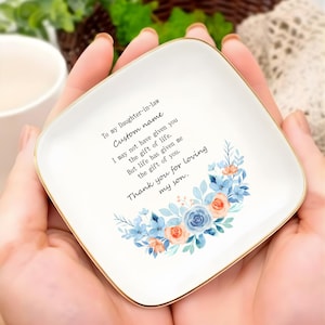 May include: A square, white ceramic dish with a gold rim, featuring a floral design in blue and orange. The dish has a printed message: "To my Daughter-in-law Custom name...Thank you for loving my son."