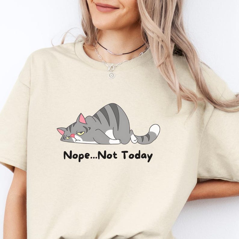 Cat themed shirt with “Nope Not Today” slogan and adorable illustrated cat, photographed indoors to emphasize comfort and trendy humor style.  A t-shirt with a cartoon gray cat lying down with the text 'Nope...Not Today' printed below the cat.