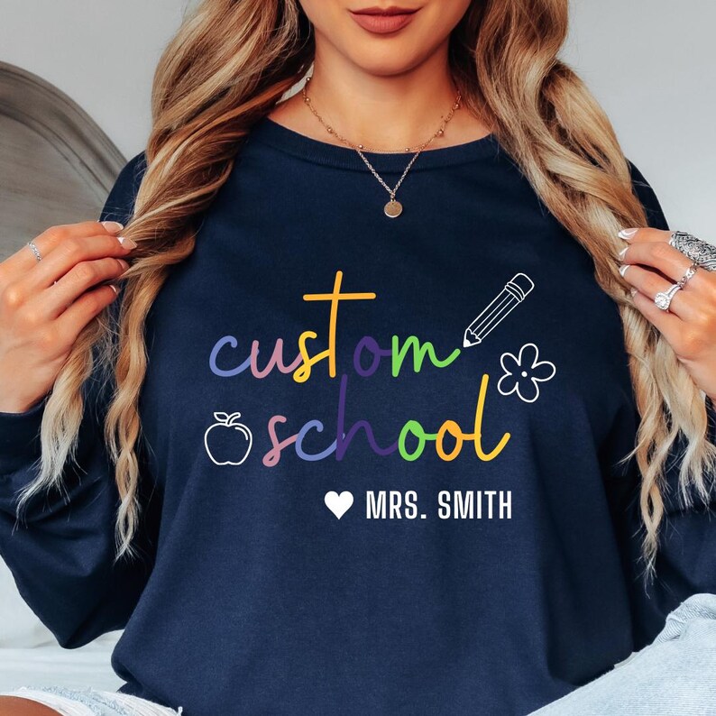 May include: A navy blue sweatshirt featuring the words "custom school" in a rainbow of colours, with an apple, pencil, and flower graphic. The text "MRS. SMITH" is below a white heart. A personalised, casual design.