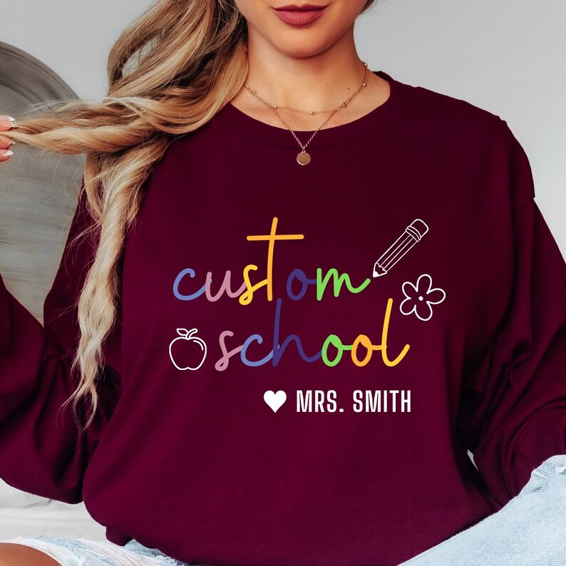 Unisex teacher sweatshirt in dark green with rainbow “custom school” design, pencil and apple graphics, and personalization option for name. Ideal for classroom wear, staff events, or appreciation gifts, combining comfort with meaningful teacher style