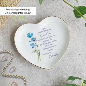 May include: A heart-shaped white ceramic dish with a gold rim, featuring blue floral artwork and personalized text. The text reads "Personalized Wedding Gift For Daughter In Law". A thoughtful gift for a daughter-in-law.