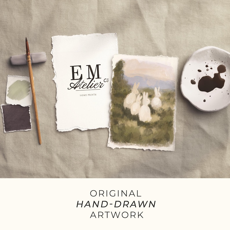 May include: A collection of original hand-drawn artwork. The image showcases a watercolour painting of white rabbits in a field, a paintbrush, paint swatches, and a small dish with dark liquid. The EM Atelier logo is also present.