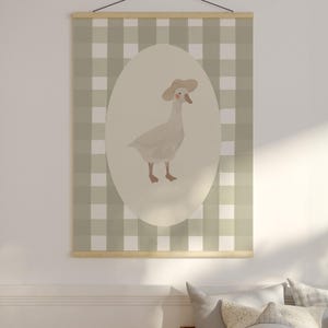 May include: A wall hanging with a watercolour illustration of a goose in a hat, set against an oval backdrop. The background is a green and white checked pattern. It is suspended by a wooden rod and black string.