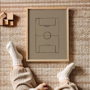 May include: A framed print featuring a minimalist soccer field design in black on a muted olive-green background. The frame is light-colored wood. The image is on a woven rug. A person's legs and feet are visible, wearing beige pants and white ribbed socks.