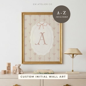 May include: A framed wall art print with a gold frame. The print features the letter "A" in a pink oval with a bow design, set against a light pink bow pattern background. The text "CUSTOM INITIAL WALL ART" is at the bottom.
