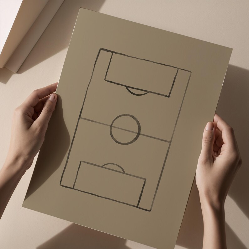 May include: A tan-colored print of a soccer field diagram, held by hands. The field is outlined in black, with the goal areas, center circle, and center line clearly marked. The paper has a matte finish.