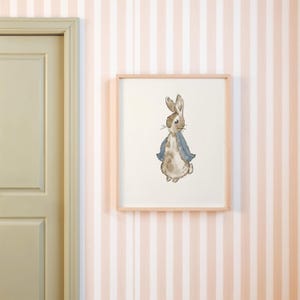 May include: Watercolor illustration of a rabbit wearing a blue jacket, framed in a light wood frame. The artwork is displayed on a wall with pink and white striped wallpaper, near a light green door.