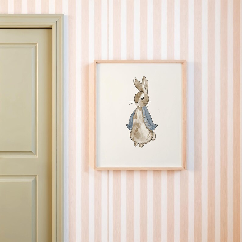 May include: Watercolor illustration of a rabbit wearing a blue jacket, framed in a light wood frame. The artwork is displayed on a wall with pink and white striped wallpaper, near a light green door.