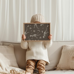 May include: A framed print featuring a repeating pattern of white dinosaur skeletons on a dark brown background. The print is held by a person wearing a cream-colored sweater, tan pants, and a knit cap. The frame is a light brown color.