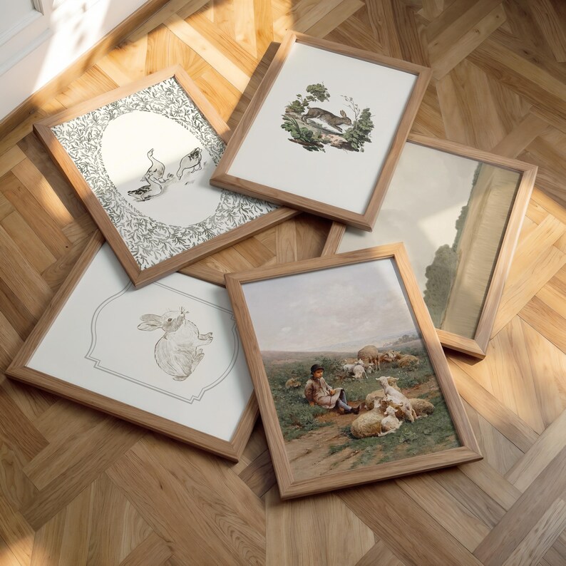May include: A selection of framed art prints with light wood frames. The prints showcase diverse illustrations, including a rabbit, a pastoral scene with sheep, and other botanical and animal-themed designs. The frames are arranged on a wooden floor.