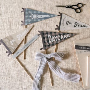 Op de afbeelding: Een verzameling decoratieve wimpels voor de eerste en laatste schooldag, waaronder "Homeschool", "8th Grade", "11th Grade" en "Daycare". De wimpels zijn in verschillende kleuren en patronen, met houten stokjes en een gestreept lint.