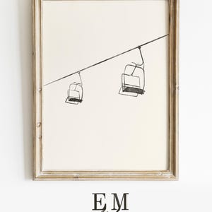 May include: A framed print with a minimalist line drawing of two ski lift chairs hanging from a cable. The artwork is in black and white, set against a cream background. The frame is light, distressed wood. The text "EM Atelier" and "DIGITAL PRINTABLE INSTANT DOWNLOAD" are at the bottom.