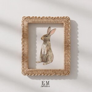 May include: A rectangular picture frame with a woven light brown rattan border containing a watercolor illustration of a brown rabbit sitting upright. The text EM ATELIER is printed in black at the bottom center of the white background.
