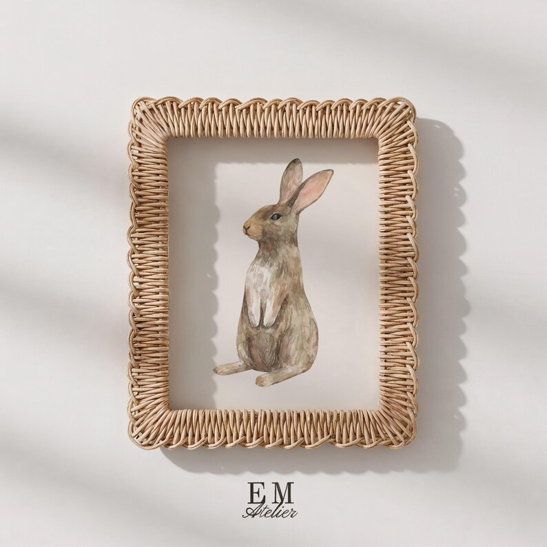 May include: A rectangular picture frame with a woven light brown rattan border containing a watercolor illustration of a brown rabbit sitting upright. The text EM ATELIER is printed in black at the bottom center of the white background.
