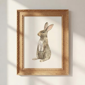 May include: A framed watercolor painting of a brown rabbit sitting upright in profile. The rabbit has long ears, a light-colored chest, and textured fur. The artwork is enclosed in a rectangular, ornate, light brown wooden frame with carved edges.
