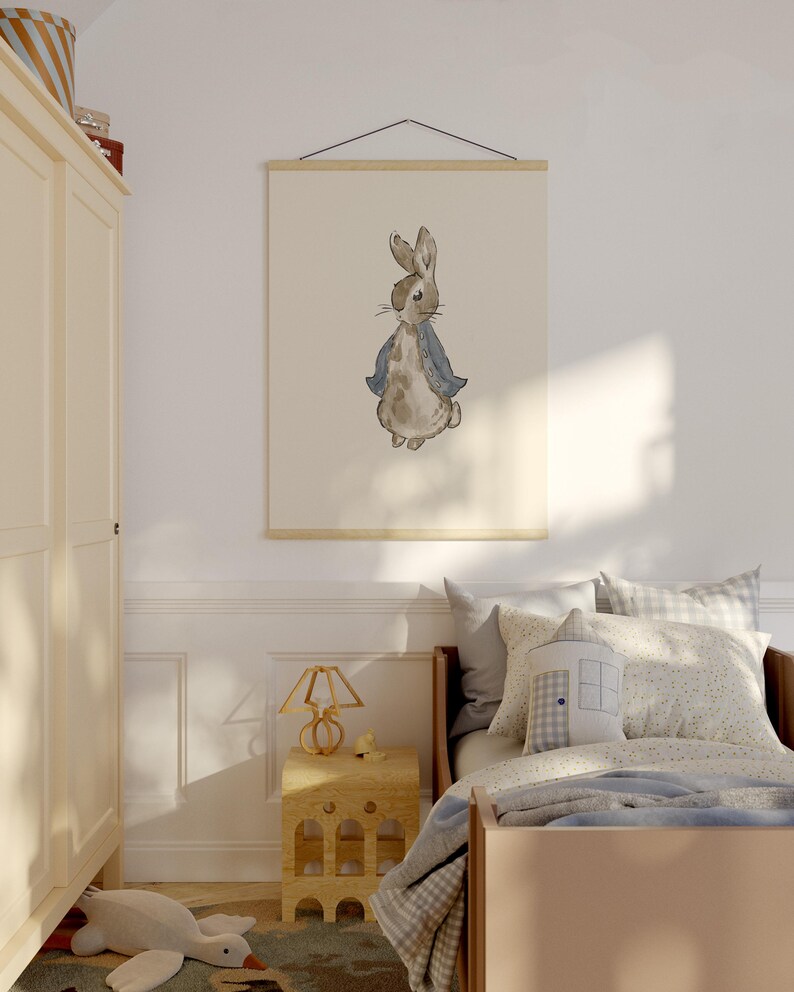 May include: Watercolor print of a rabbit wearing a blue jacket, hanging on a wall in a child's bedroom. The artwork is displayed on a wooden frame. The room features a bed, nightstand, and a wardrobe.