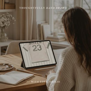 May include: A digital illustration of a baseball jersey with the name "SMITH" and the number "23" displayed on a tablet. The text "THOUGHTFULLY HAND DRAWN" and "WITH LOVE, EMILIE" are also visible. A paintbrush, palette, and notebook are on the table.