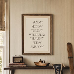 May include: A framed print displays the days of the week in a minimalist style. The text is in a simple font, set against a cream background, and framed in dark wood. The print hangs above a shelf with a vintage radio, books, and a wooden boat model.