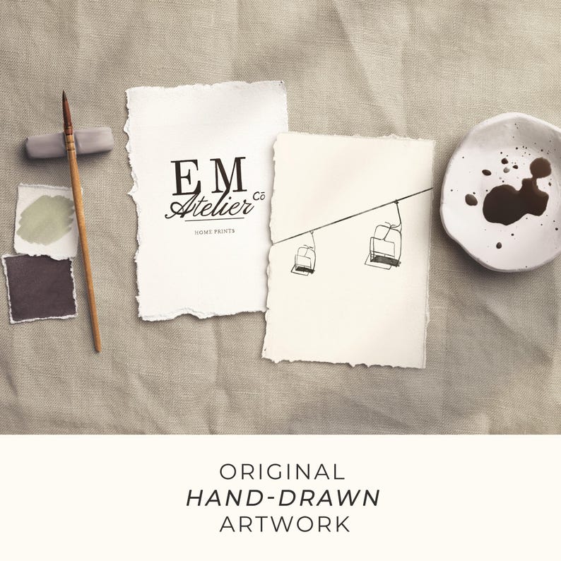 May include: Hand-drawn artwork featuring two pieces of paper with torn edges. One displays the text "EM Atelier Co HOME PRINTS" in black, the other a sketch of a ski lift. A paintbrush, paint swatches, and a small dish with brown paint are also present.