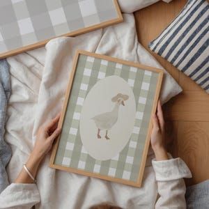 May include: A framed print with a cartoon goose wearing a hat, set against a green and white gingham background. The goose is inside an oval shape. The frame is made of light-coloured wood. Additional framed prints with a similar pattern are visible.