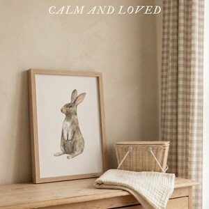 May include: A framed watercolor print of a brown rabbit in a light wood frame. The text above reads: CREATED TO MAKE YOUR SPACE FEEL CALM AND LOVED. Below the frame, a small woven basket and a folded cream-colored fabric cloth are visible.