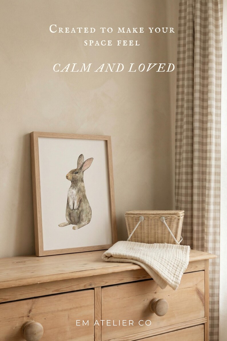 May include: A framed watercolor print of a brown rabbit in a light wood frame. The text above reads: CREATED TO MAKE YOUR SPACE FEEL CALM AND LOVED. Below the frame, a small woven basket and a folded cream-colored fabric cloth are visible.