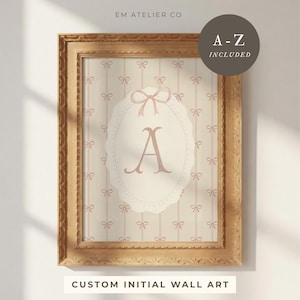 May include: A framed custom initial wall art print. The print features a light pink letter "A" inside an oval shape with a bow design. The background has a striped pattern with small bows. The frame is gold and the text "A-Z INCLUDED" is in a circle.