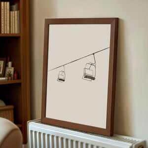 May include: A framed art print showing a minimalist sketch of two ski lift chairs hanging from a cable. The artwork is in black on a cream background, set within a dark brown wooden frame. The print is leaning against a wall.