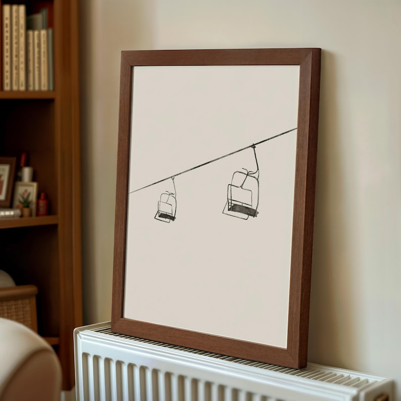 May include: A framed art print showing a minimalist sketch of two ski lift chairs hanging from a cable. The artwork is in black on a cream background, set within a dark brown wooden frame. The print is leaning against a wall.