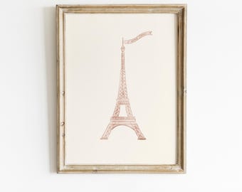 Può includere: Una stampa incorniciata con un'illustrazione ad acquerello della Torre Eiffel in una delicata tonalità rosa. La torre ha uno stendardo in cima. La stampa è su uno sfondo color crema e incorniciata in una cornice di legno dorato.