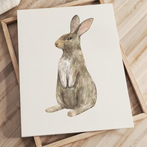 May include: A rectangular wall art print featuring a watercolor illustration of a brown rabbit sitting upright. The rabbit is depicted in profile, facing left, with a white chest and soft brown fur tones against a plain, off-white background.