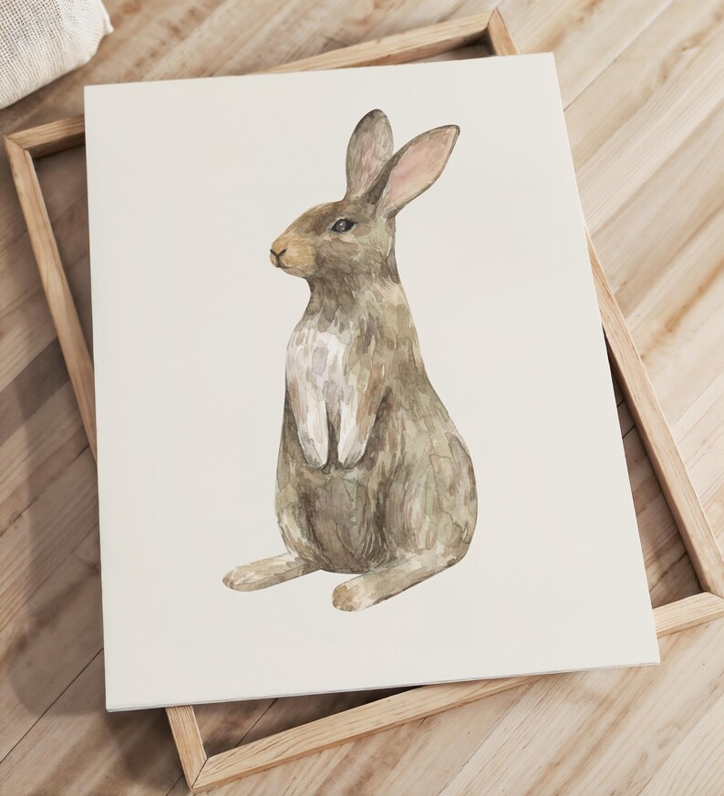 May include: A rectangular wall art print featuring a watercolor illustration of a brown rabbit sitting upright. The rabbit is depicted in profile, facing left, with a white chest and soft brown fur tones against a plain, off-white background.