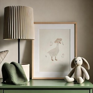 Peut inclure: Une impression encadr&eacute;e d'une oie portant un chapeau, avec un fond beige et un cadre en bois. Une lampe beige avec un abat-jour pliss&eacute;, une couverture en tricot vert et un lapin en peluche sont sur une surface verte.