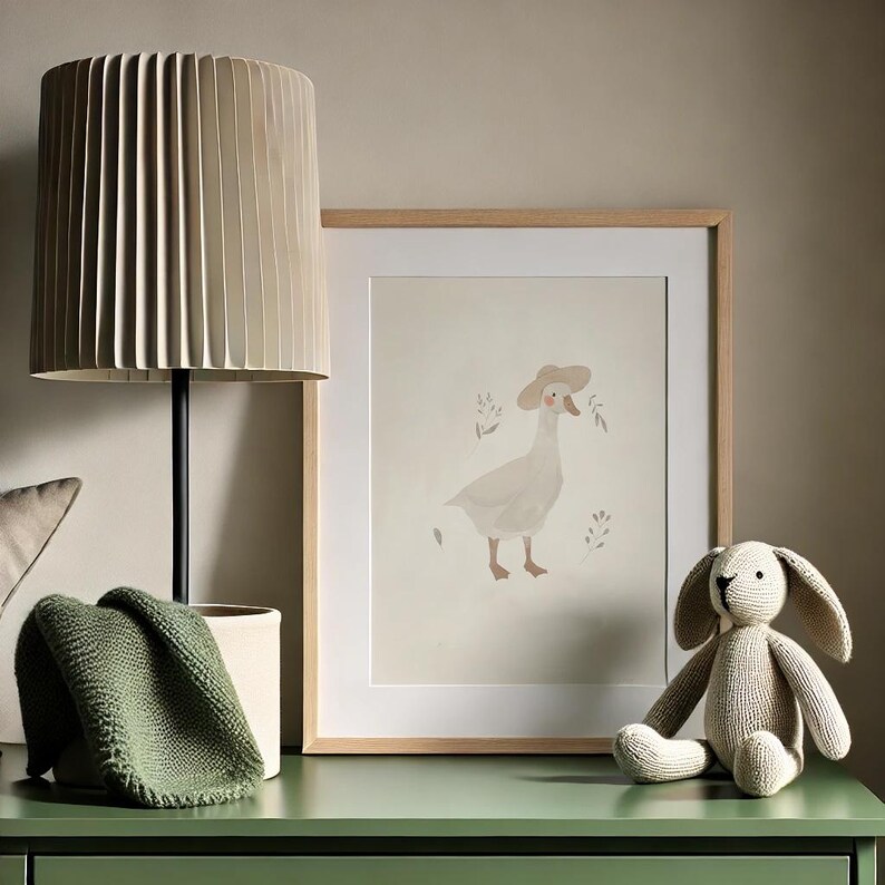 Peut inclure: Une impression encadr&eacute;e d'une oie portant un chapeau, avec un fond beige et un cadre en bois. Une lampe beige avec un abat-jour pliss&eacute;, une couverture en tricot vert et un lapin en peluche sont sur une surface verte.