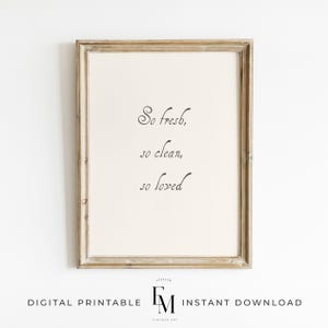 May include: A printable wall art print with a white background and a black cursive font that reads "So fresh, so clean, so loved". The print is framed in a light brown wood frame.
