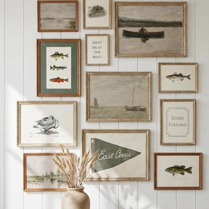 May include: A collection of framed art prints with a coastal theme, including illustrations of fish, boats, and landscapes. The frames are a warm brown, and the artwork is arranged on a white paneled wall. A vase with dried wheat and a wooden table complete the scene.