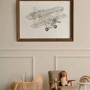 May include: A framed black and white illustration of a vintage airplane, hanging on a white wall with wainscoting. A wicker baby carriage and a plush toy are in the foreground. The text "TIMELESS PRINTS FOR KIDS" is at the top of the image. The text "@EMATELIERCO" is at the bottom of the image.