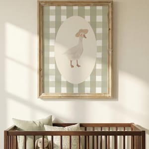 May include: A framed print with a watercolour illustration of a goose wearing a hat, set against a green and white gingham background. The artwork is in a light wooden frame, above a wooden cot with pillows and a stuffed toy.