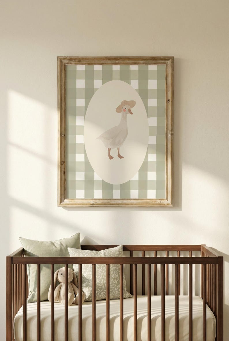 May include: A framed print with a watercolour illustration of a goose wearing a hat, set against a green and white gingham background. The artwork is in a light wooden frame, above a wooden cot with pillows and a stuffed toy.
