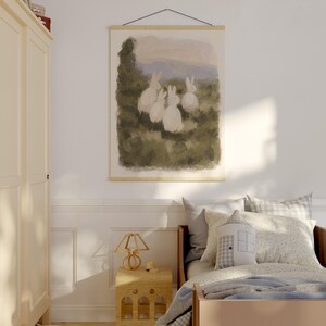 May include: A framed print depicting a watercolour scene of five white rabbits in a field. The artwork hangs above a child's bed with a wooden frame, pillows, and a patterned blanket. A wooden bedside table and lamp are also visible.
