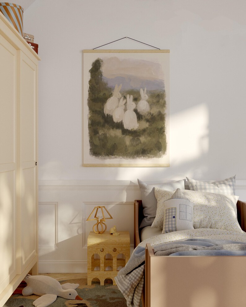 May include: A framed print depicting a watercolour scene of five white rabbits in a field. The artwork hangs above a child's bed with a wooden frame, pillows, and a patterned blanket. A wooden bedside table and lamp are also visible.