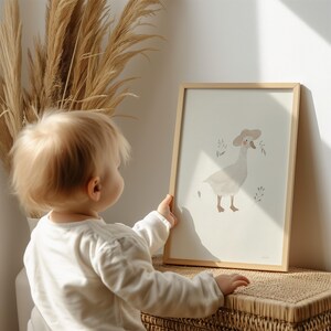 Peut inclure: Une affiche encadr&eacute;e avec une illustration d'oie portant un chapeau, avec des brins de feuilles. L'&oelig;uvre est dans des tons doux et sourds, dans un cadre en bois clair. L'affiche est tenue par un enfant.