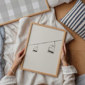 May include: A framed print with a minimalist line drawing of two ski lift chairs suspended from a cable. The artwork has a light beige background and a natural wood frame. Another framed print with a grey and white checkered pattern is also visible.