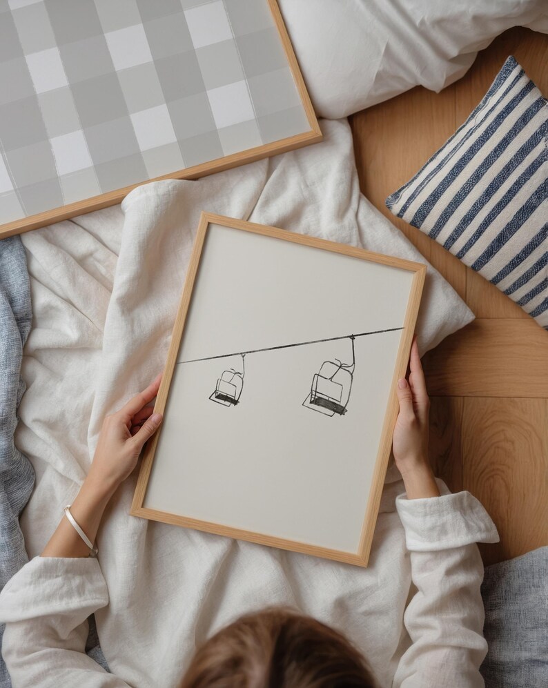 May include: A framed print with a minimalist line drawing of two ski lift chairs suspended from a cable. The artwork has a light beige background and a natural wood frame. Another framed print with a grey and white checkered pattern is also visible.
